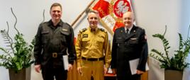 Na zdjęciu widać trzech strażaków. W środku stoi komendant główny PSP, w tle sztandar z godłem RP.