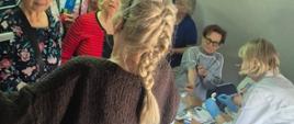grupa osób stoi przy stoliku z ulotkami a przed nimi stoi pracownica promocji zdrowia obok stoi pracownica przychodni rodzinnej ubrana w biały fartuch i przeprowadza badania cukru we krwi oraz badania ciśnienia tętniczego 