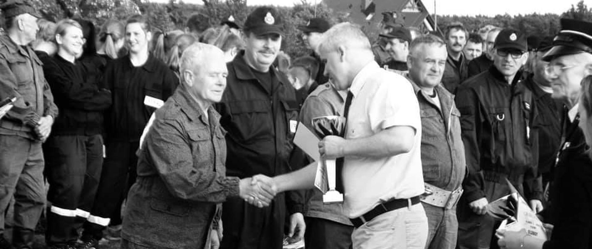 Druh Zdzisław Hałuszczak odbiera puchar z rąk Pana Jacka Kolesińskiego, podczas zbiórki końcowej na zawodach sportowo pożarniczych. Stoi pierwszy z lewej, po jego prawej stronie oraz w tle przedstawiciele pozostałych jednostek OSP. Zdjęcie czarno-białe.