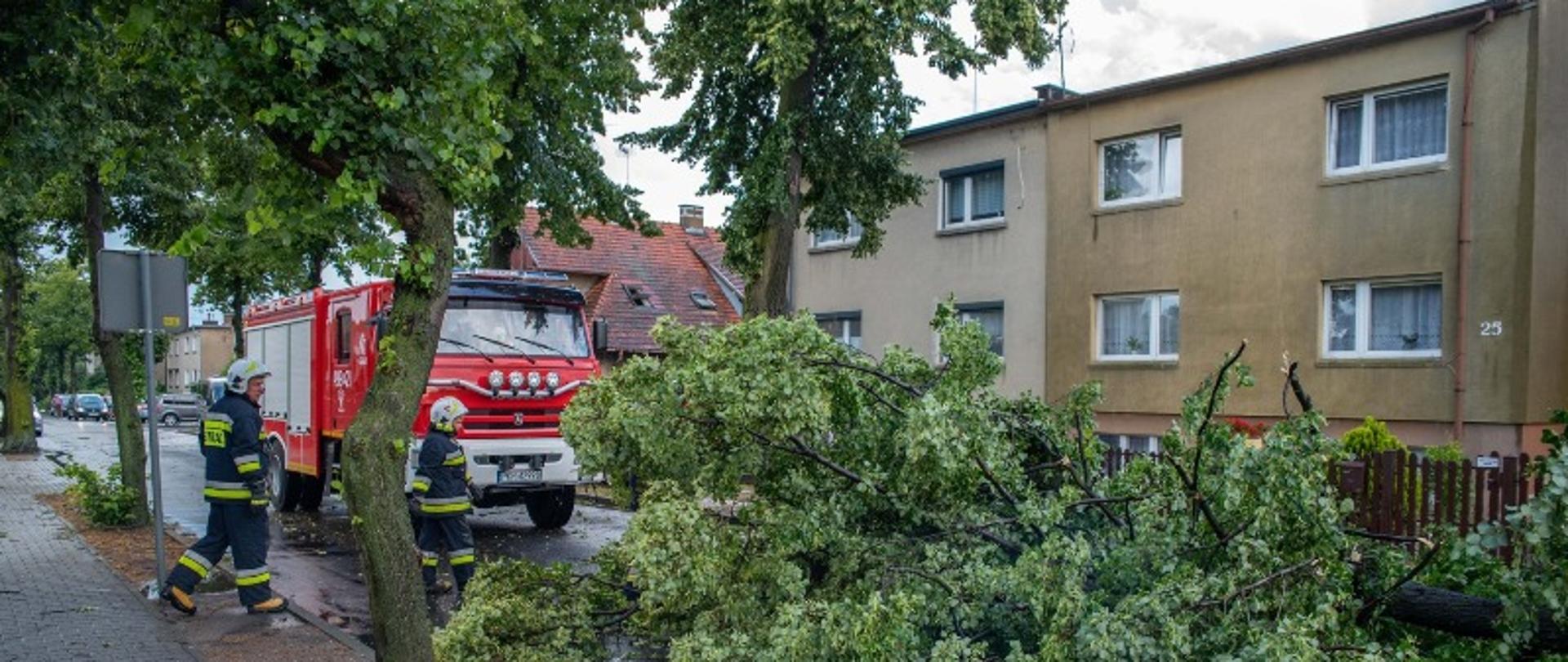 Na drodze zalega konar drzewa, który blokuje ruch pojazdów. Na miejsce dotarł samochód gaśniczy z jednostki OSP Koźmin WIelkopolski. Przed pojazdem widać dwóch strażaków w białych hełmach i ubraniach bojowych, kórzy przygotowują się do usunięcia wiatrołomu. Pogoda deszczowa i wietrzna. Obok drogi znajdują się budynki jednorodzinne w zabudowanie bliźniaczej.