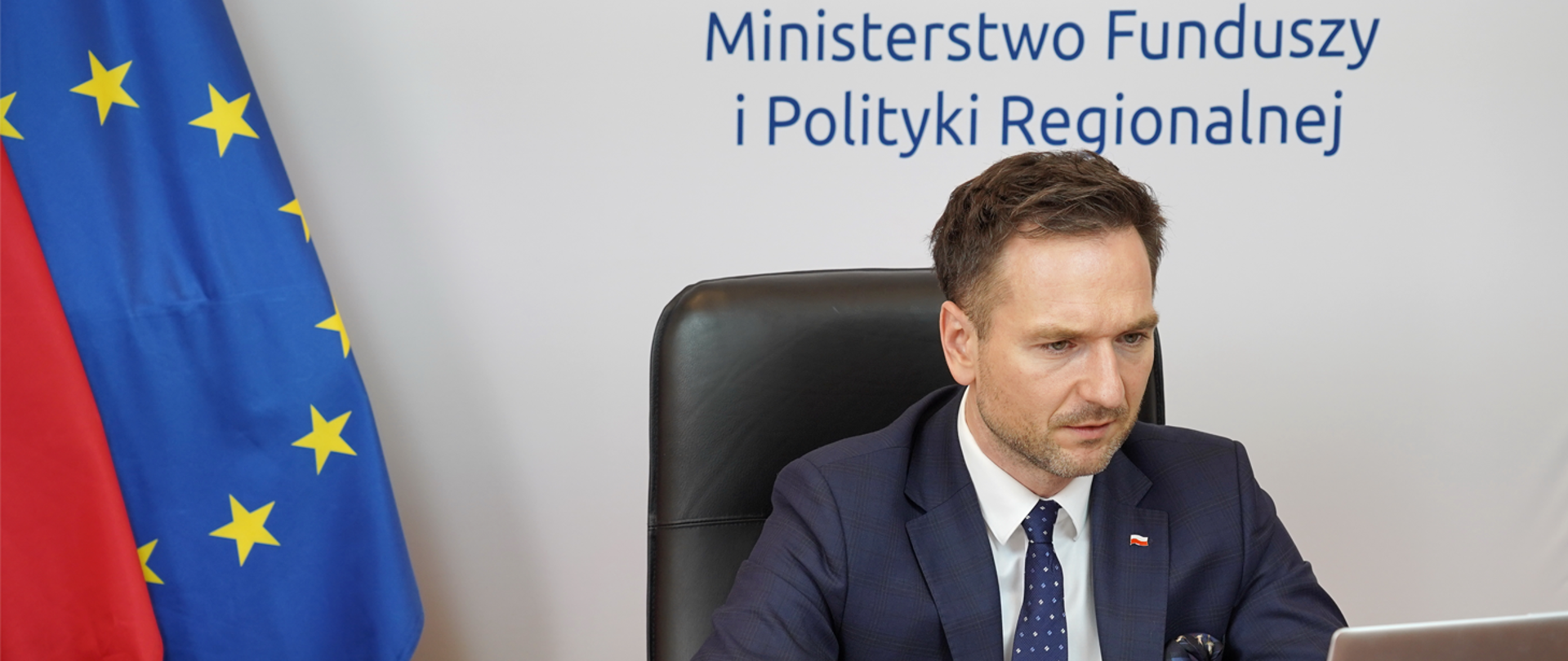 Wiceminister Waldemar Buda siedzi w fotelu i patrzy na ekran laptopa. Za nim flagi PL i UR oraz napis na ściance: Ministerstwo Funduszy i Polityki Regionalnej.