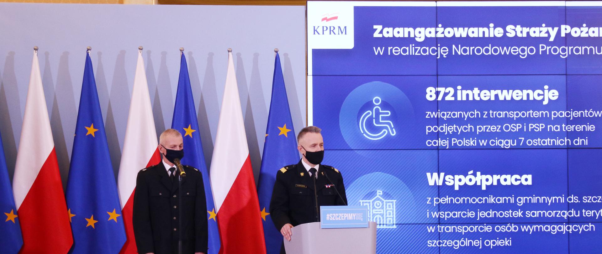 Komendant Główny Państwowej Straży Pożarnej nadbryg. Andrzej Bartkowiak oraz druh Andrzej Matczak podczas konferencji prasowej. Z tyłu biało-czerwone flagi oraz flagi Unii Europejskiej oraz ekran prezentacyjny