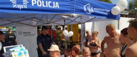 Na zdjęciu policjant na stanowisku edukacyjnym pod namiotem prowadzi prelekcje dla zainteresowanych