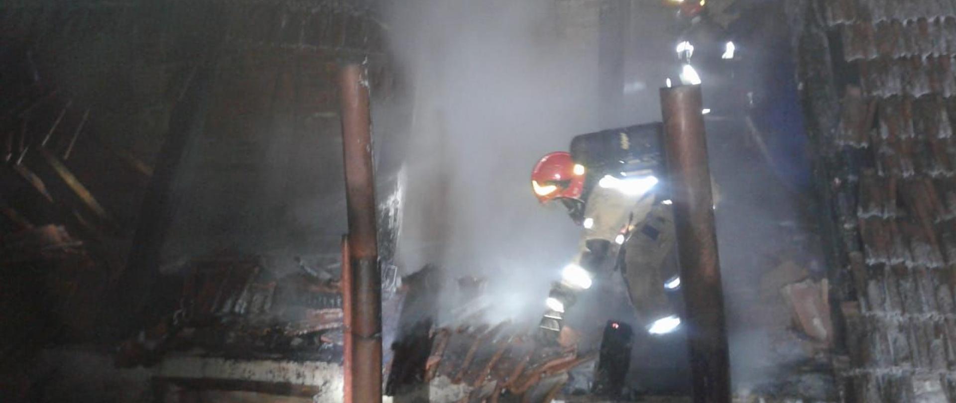 Pożar połaci dach budynku gospodarczego o wielkości piętnastu metrów kwadratowych. Centralnie widać strażaka podczas dogaszania konstrukcji dachu. 