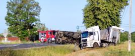 Na zdjęciu widoczna jest przewrócona biała ciężarówka z naczepą, leży na boku w rowie, a na drodze obok stoi czerwony wóz strażacki, co wskazuje na miejsce wypadku drogowego. W tle widać jakieś zabudowania otoczone czerwonym murem i linię energetyczną.