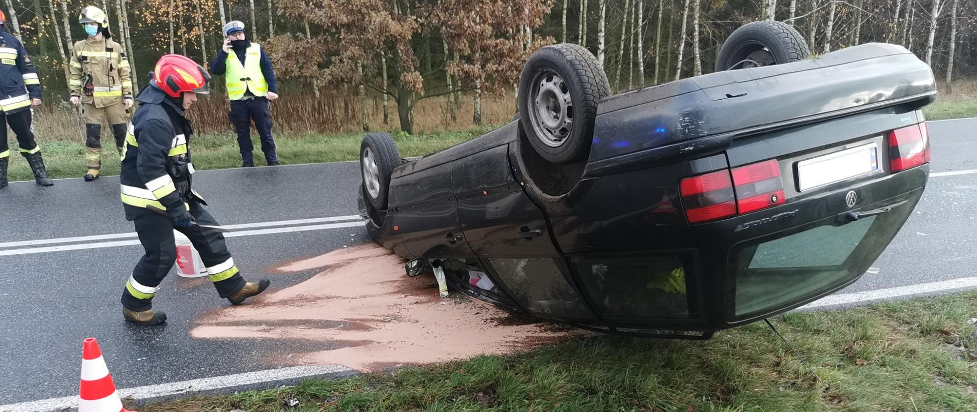 Na zdjęciu widnieje czarny pojazd znajdujący się na dachu. W lewej części zdjęcia widzimy strażaka ubranego w ubranie specjalne oraz hełm, który rozsypuje pomarańczowy proszek na asfalt. Obok niego znajduje się pachołek. Pomiędzy pojazdem, a strażakiem asfalt jest wysypany pomarańczowym proszkiem. W tle znajduje się dwóch strażaków oraz policjant. W oddali widzimy drzewa.