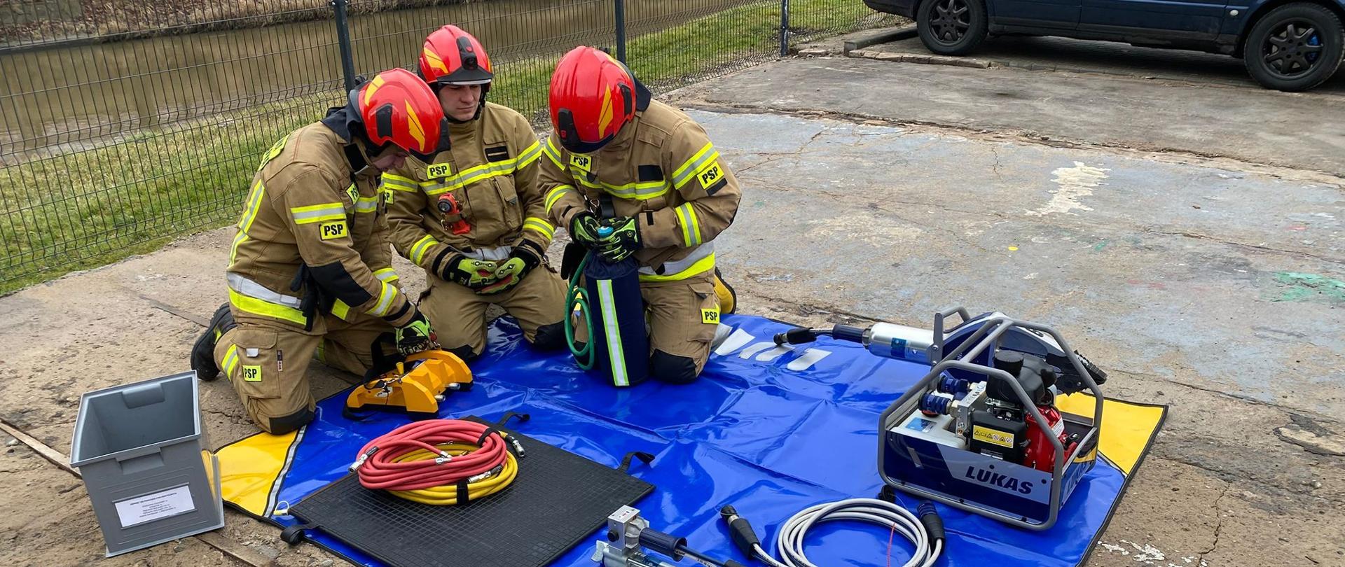 Trzech strażaków w mundurach bojowych i czerwonych hełmach, siedzą przy niebieskiej macie, na macie sprzęt hydrauliczny, trwają ćwiczenia.