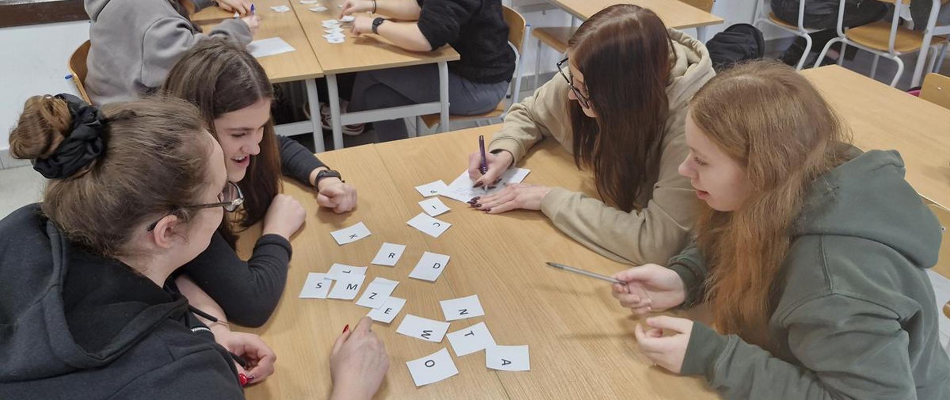 Grupa młodzieży siedzi przy stolikach w sali lekcyjnej i wspólnie układa słowa z białych kartoników z literami.