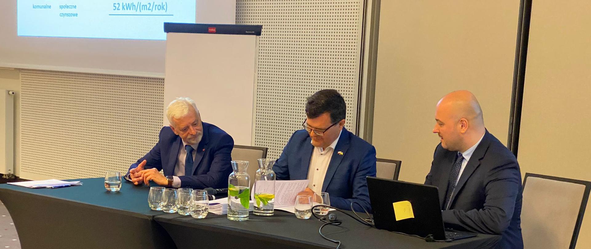 Wiceminister Piotr Uściński podpisuje pismo siedząc za stołem, w tle ekran z prezentacją