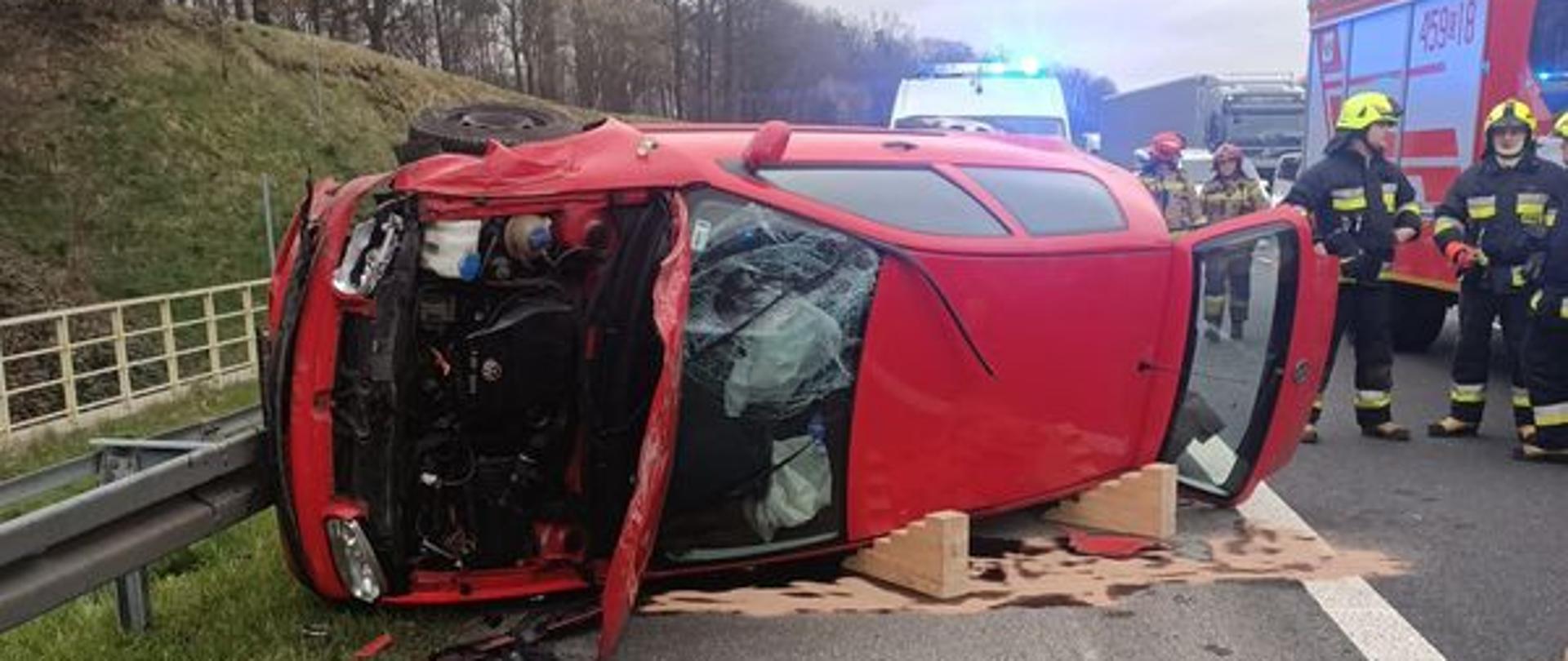 Na zdjęciu znajduje się wrak samochodu przewrócony na boku, obok widać samochód pożarniczy oraz strażaków, w tle radiowóz 