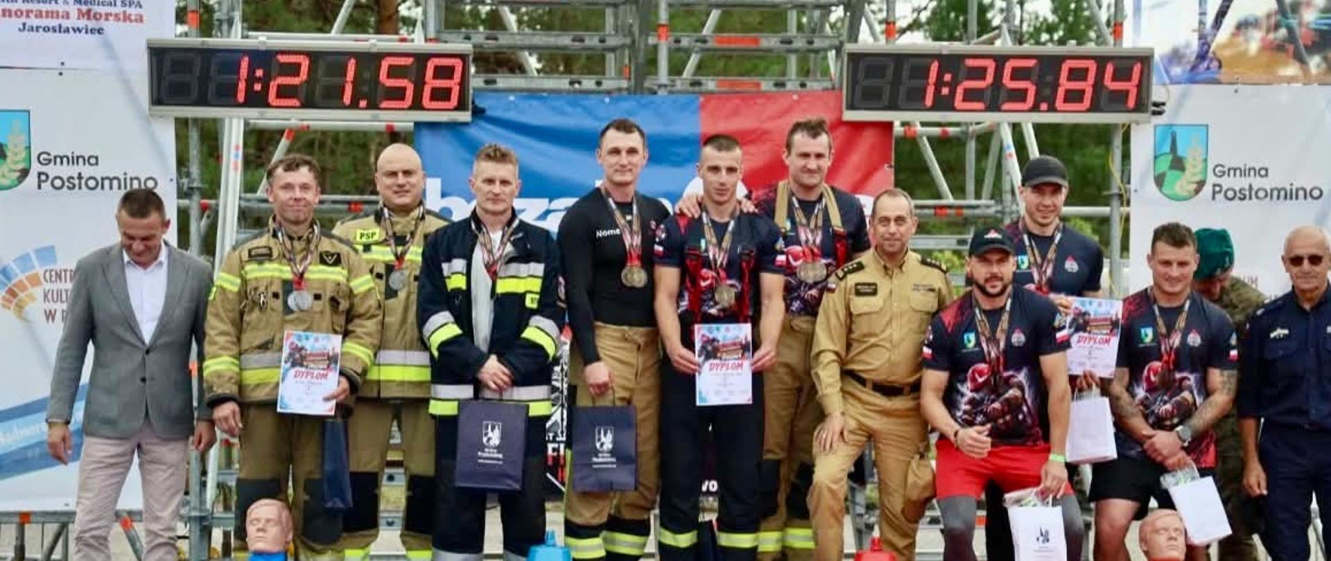 Zdjęcie przedstawia grupę strażaków w umundurowaniu stojących na podium podczas zawodów. Na pierwszym miejscu znajduje się drużyna z czasem 1:25,84, na drugim z czasem 1:21,59, a na trzecim miejscu kolejna drużyna. Uczestnicy trzymają dyplomy i puchary, a przed podium widoczne są elementy wyposażenia strażackiego. W tle znajdują się banery sponsorów i dekoracje związane z zawodami.