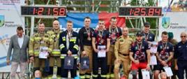 Zdjęcie przedstawia grupę strażaków w umundurowaniu stojących na podium podczas zawodów. Na pierwszym miejscu znajduje się drużyna z czasem 1:25,84, na drugim z czasem 1:21,59, a na trzecim miejscu kolejna drużyna. Uczestnicy trzymają dyplomy i puchary, a przed podium widoczne są elementy wyposażenia strażackiego. W tle znajdują się banery sponsorów i dekoracje związane z zawodami