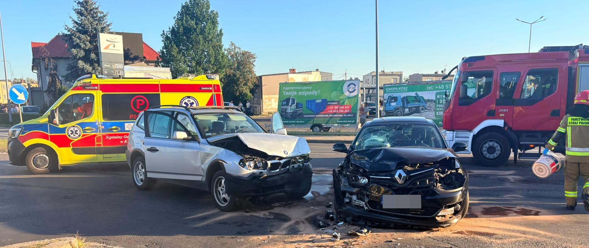 Na zdjęciu widać miejsce kolizji drogowej. Na pierwszym planie znajduje się mocno uszkodzony czarny samochód osobowy z rozbitą przednią częścią. Za nim stoi uszkodzony srebrny samochód. W tle widoczna jest karetka pogotowia z włączonymi niebieskimi światłami oraz ratownik. Na jezdni leżą fragmenty uszkodzonych pojazdów i widoczne są plamy płynów. Zdarzenie ma miejsce na skrzyżowaniu w zabudowanym obszarze, w słoneczny dzień.