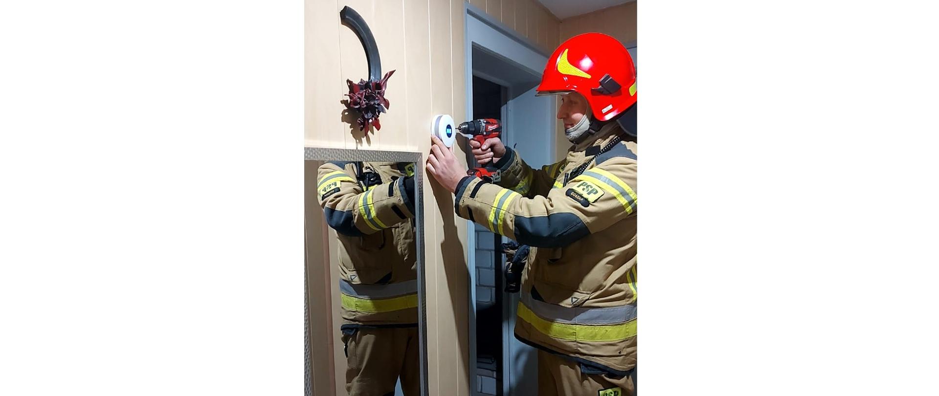 Zdjęcie przedstawia strażaka z Komendy Powiatowej Państwowej Straży Pożarnej w Gołdapi, który montuje czujkę tlenku węgla i dymu w mieszkaniu gdzie wykryto obecność czadu