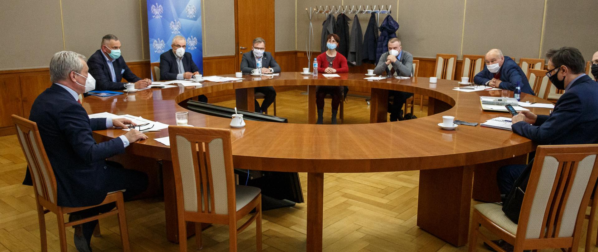 Na zdjęciu 9 osób zasiadających przy okrągłym stole na sali im. Braci Kowalczyków w urzędzie. Po lewej stronie wojewoda opolski, prowadzący spotkanie. 