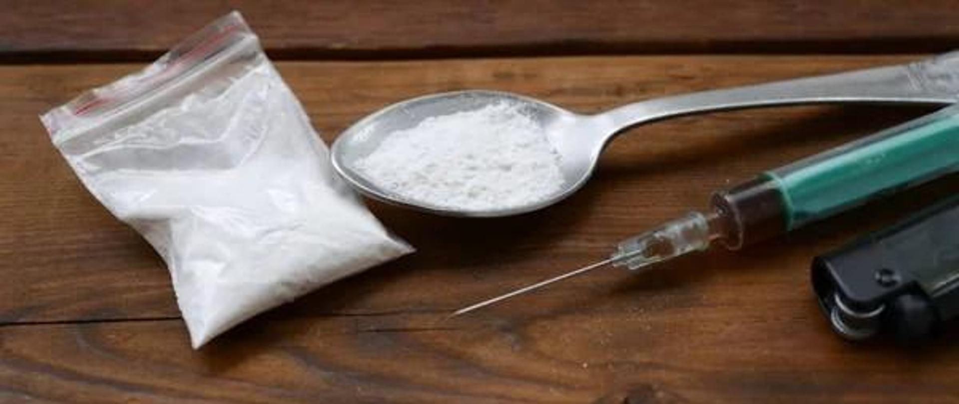 Grafika przedstawia torebkę oraz łyżkę z białym proszkiem, a także strzykawek z brązowym płynem