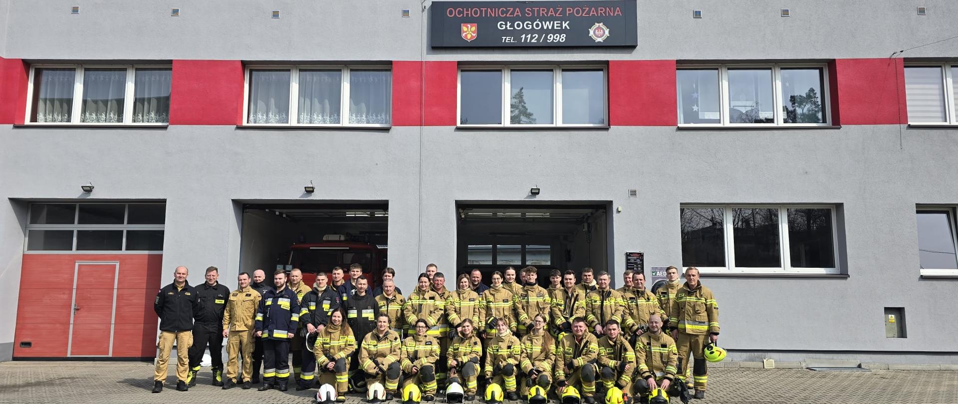 Zdjęcie przedstawia grupę strażaków w mundurach stojących przed budynkiem remizy OSP