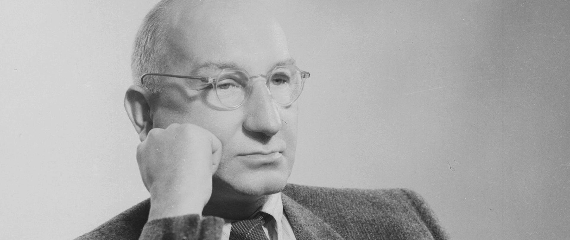 Wincenty Rzymowski - portret łysiejącego mężczyzny w okularach, siedzącego na fotelu, podpierającego pięścią twarz. Ciemna marynarka, ciemny krawat, biała koszula.