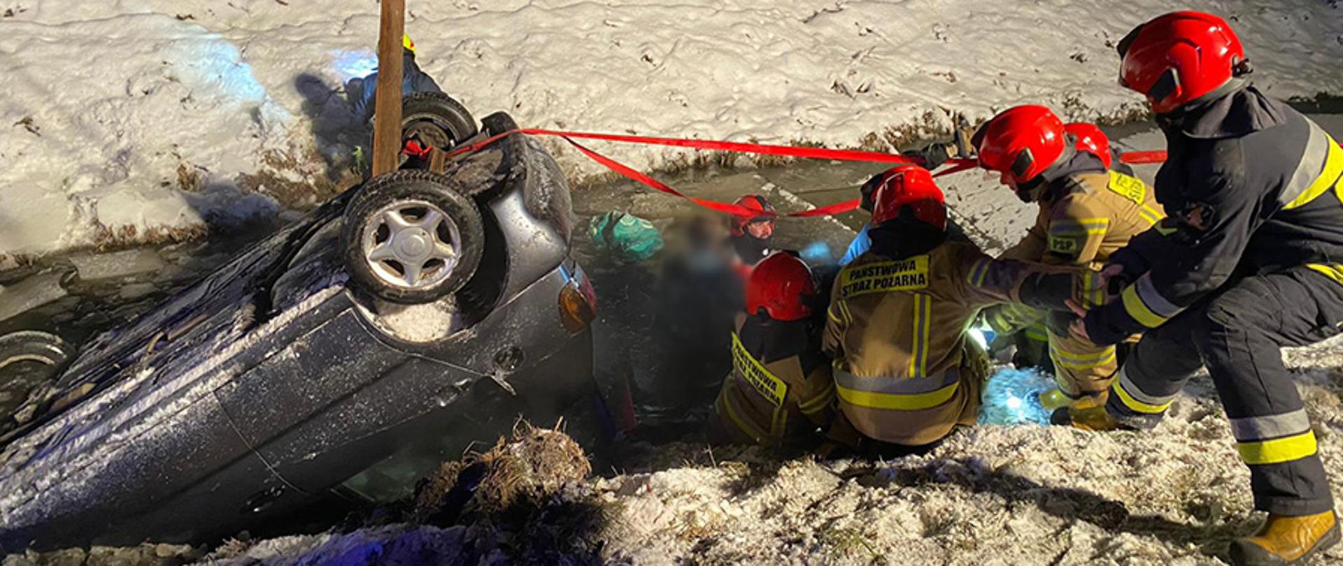 Dwóch strażaków ubranych w wodery ewakuuje osoby poszkodowane z auta, które wpadło do przydrożnego rowu wypełnionego wodą. Strażacy ubrani w wodery przekazują osobę poszkodowaną ratownikom znajdującym się na londzie.