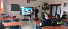 Zdjęcie przedstawia strażaków w sali szkoleniowej, którzy słuchają policjanta prowadzącego szkolenie