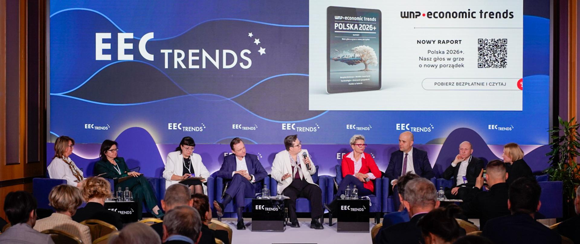 Widok na scenę konferencji EEC Trends z perspektywy widowni (widoczne plecy osób siedzących w sali). Na scenie siedzi dziewięcioro panelistów. Na głównym ekranie nad nimi wyświetlana jest grafika promująca nowy raport „WNP Economic Trends: Polska 2026+” wraz z kodem QR.