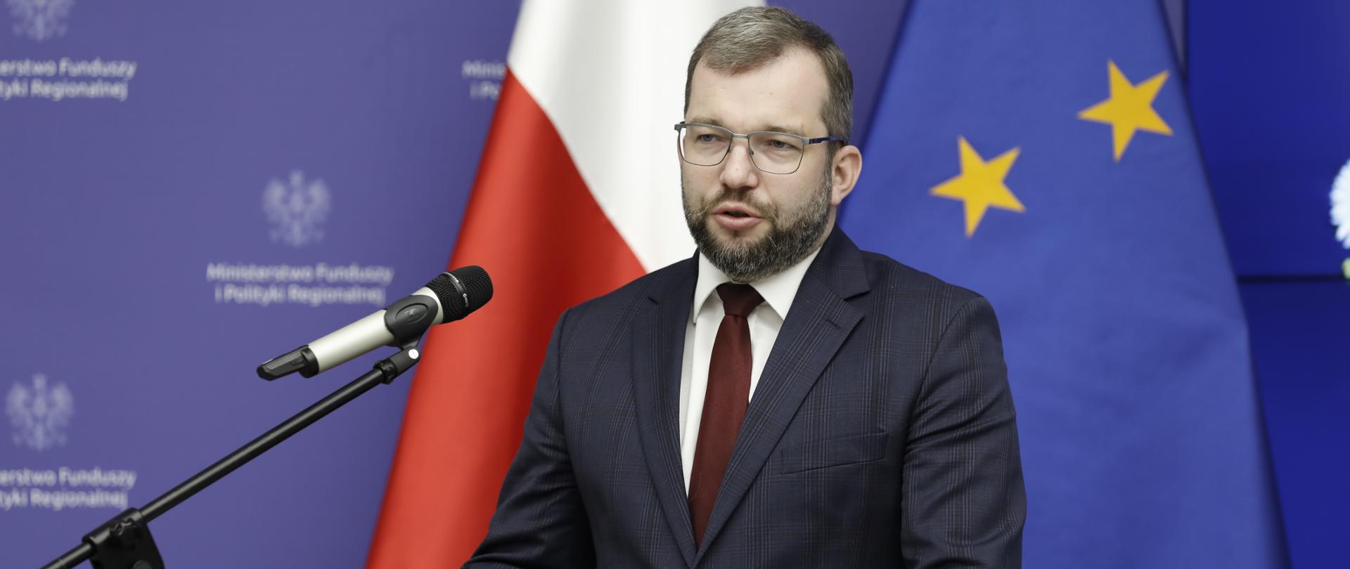 Minister Grzegorz Puda stoi przy pulpicie i mówi do ludzi zgromadzonych na sali.