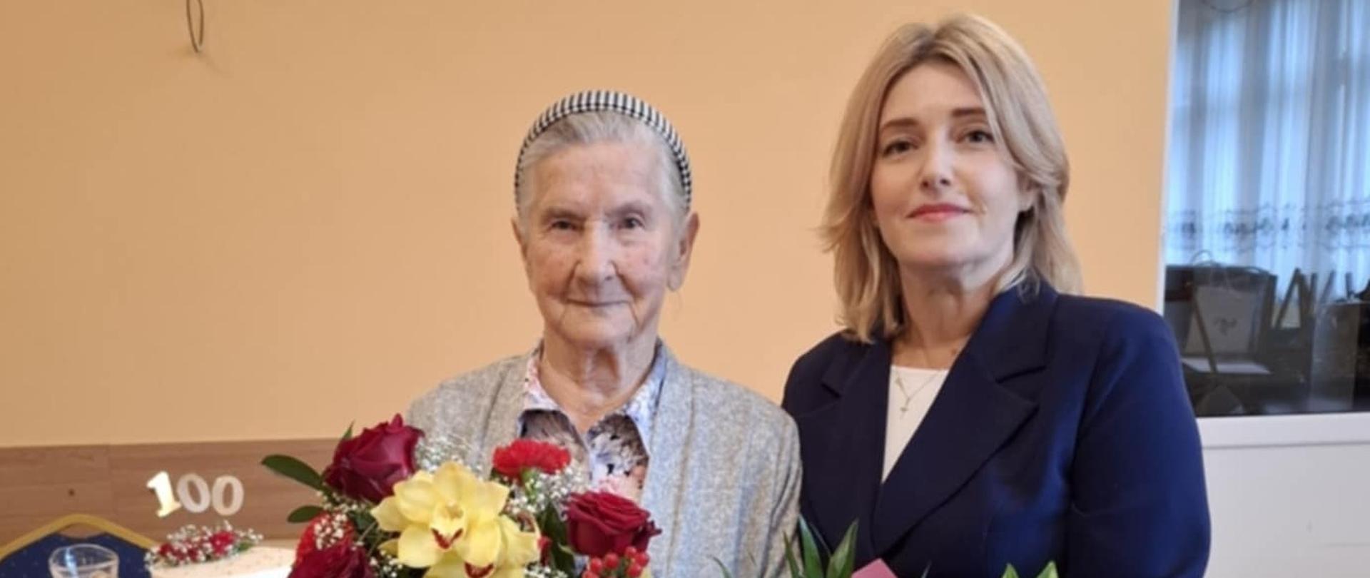 Starsza kobieta w jasnym stroju z bukietem czerwonych i żółtych kwiatów pozuje do zdjęcia stojąc obok młodej kobiety w granatowym garniturze trzymającej bukiet różowych kwiatów. W tle stół na którym stoi tort z ozdobą w kształcie liczby 100.