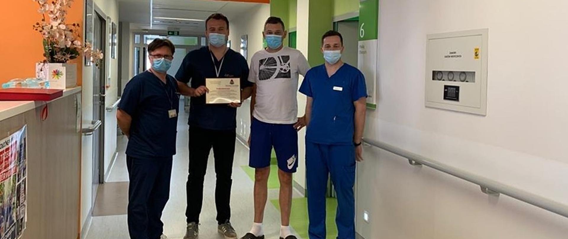 Korytarz w szpitalu. Czterech mężczyzn stoi na środku. Trzech z nich ubranych w granatowe uniformy medycznne i maseczki na twarzach. Drugi od prawej mężczyzna ubrany w jasną koszulkę i niebieskie krótkie spodenki ( Tomasz Kożuch). Pamiątkowe zdjęcie po przekazaniu podziękowań.