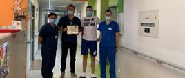 Korytarz w szpitalu. Czterech mężczyzn stoi na środku. Trzech z nich ubranych w granatowe uniformy medycznne i maseczki na twarzach. Drugi od prawej mężczyzna ubrany w jasną koszulkę i niebieskie krótkie spodenki ( Tomasz Kożuch). Pamiątkowe zdjęcie po przekazaniu podziękowań.