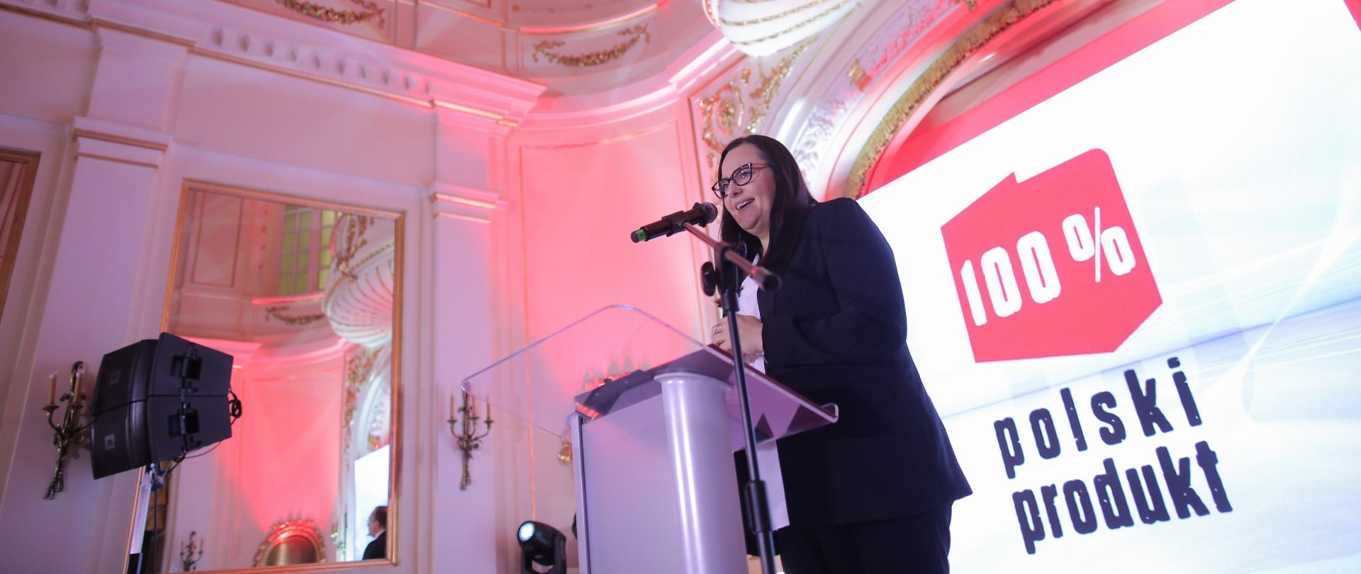 W sali oświetlonej na fioletowo, na tle ścianki z napisem 100% Polski Produkt, przemawia Małgorzata Jarosińska-Jedynak.