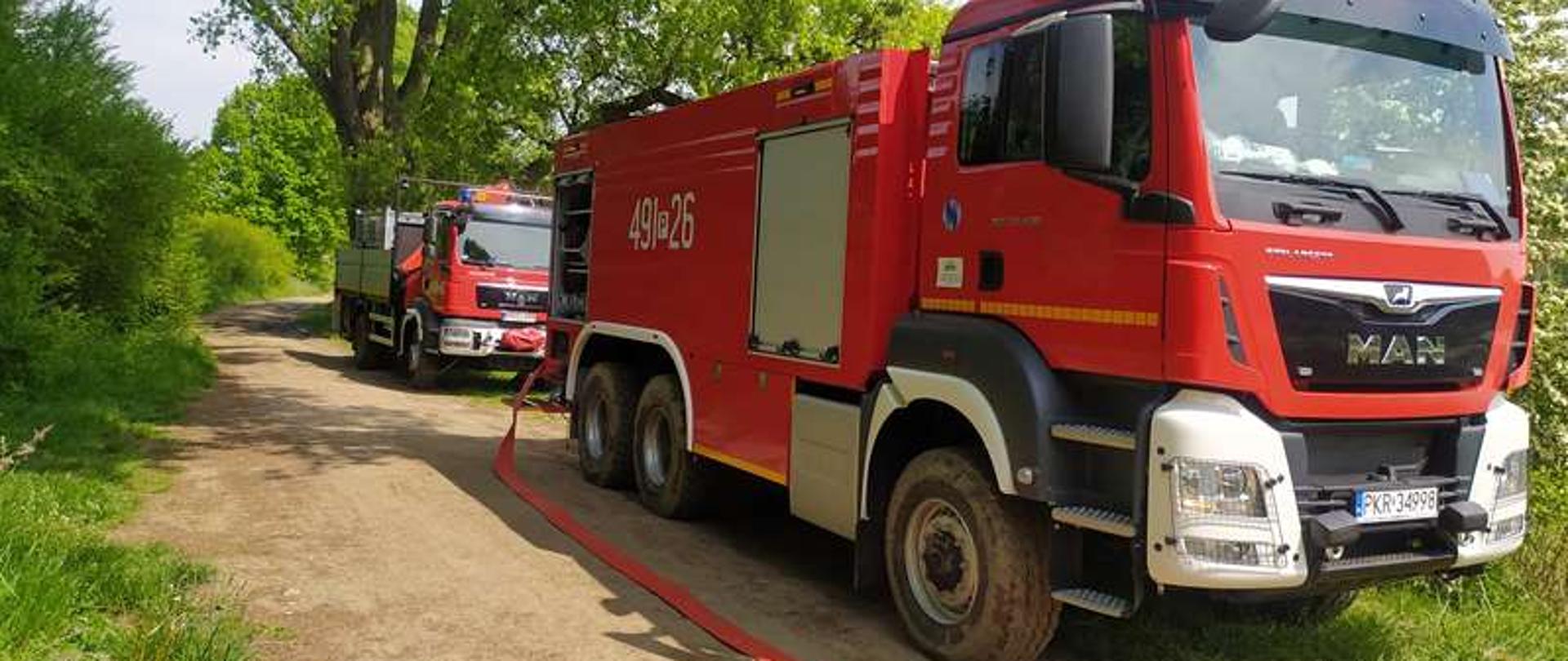 Na drodze w kompleksie leśnym stoją dwa samochody pożarnicze. Czerwony samochód gaśniczy trzyosiowy zasila linie gaśniczą. Za pojazdem gaśniczym stoi samochód wężowy na którym znajdują się odcinki węża do budowania linii. 