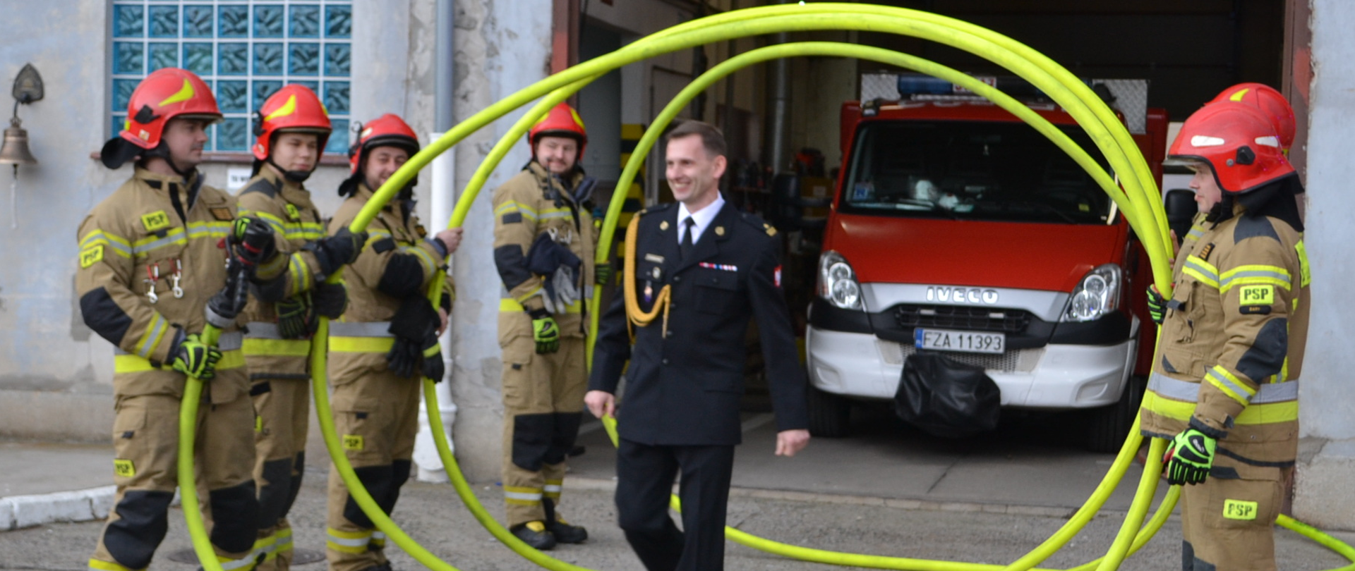 Strażak przechodzi przez szpaler tworzony przez strażaków w mundurach bokowych i czerwonych hełmach. Strażacy trzymają wąż strażacki wypełniony wodą zwinięty w spiralę.