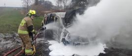 na zdjęciu spalony całkowicie samochód z otwartą maską silnika - pod samochodem plama biała piany gaśniczej obok niego strażak podaje prąd piany z żółtego pojemnika na palący się pojazd widać kłęby białego dymu w oddali pole uprawne oraz drzewa