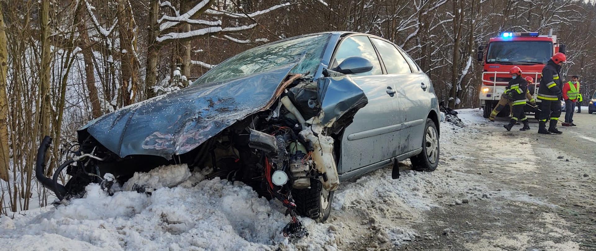 Samochód z rozbitym przodem stoi na zaśnieżonym poboczu. W tle pracujący strażacy oraz samochód pożarniczy.