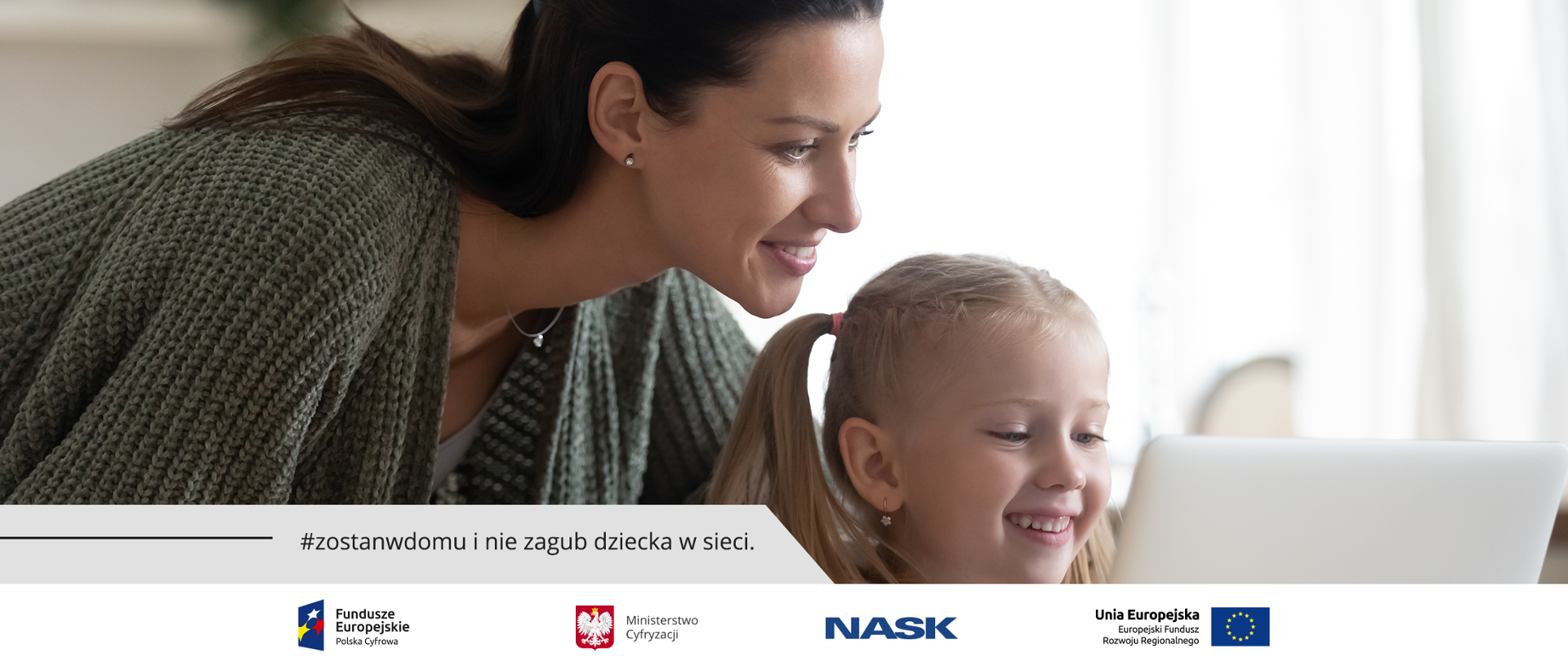 Uśmiechnięta dziewczynka siedzi przed laptopem. Za nią stoi jej mama i tez patrzy w monitor