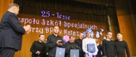 Minister Czarnek stoi obok grupy ludzi i klaszcze. Za nimi złota kotara a na niej niebieski napis 25-lecie Zespołu Szkól specjalnych przy DPS w Matczynie.