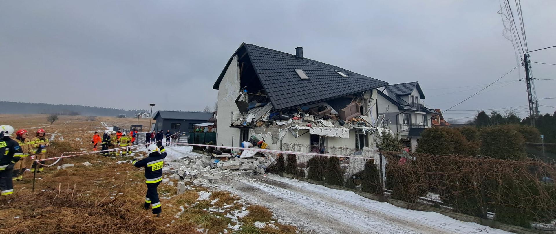 Fotografia przedstawia budynek zniszczony wybuchem, wokół którego strażacy wyznaczają strefę niebezpieczną ograniczając dostęp osób postronnych.