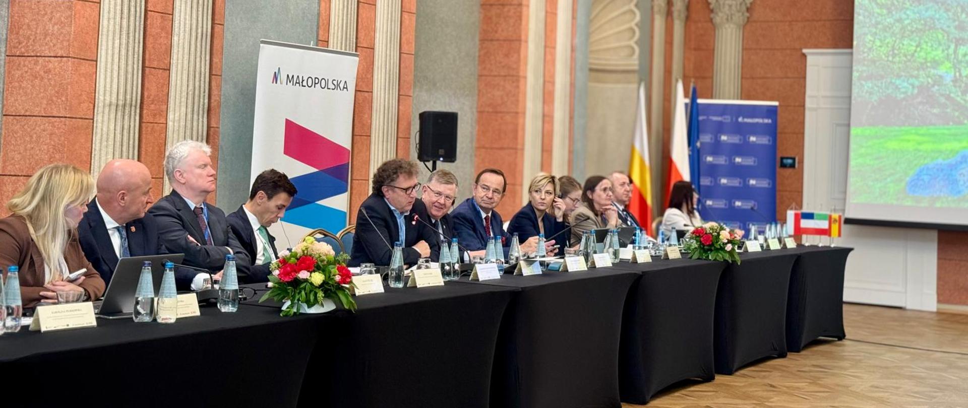 Dwanaście osób siedzi obok siebie z lewej strony przy stole. Wśród nich w środku siedzi wiceminister Jacek Karnowski i mówi do mikrofonu.