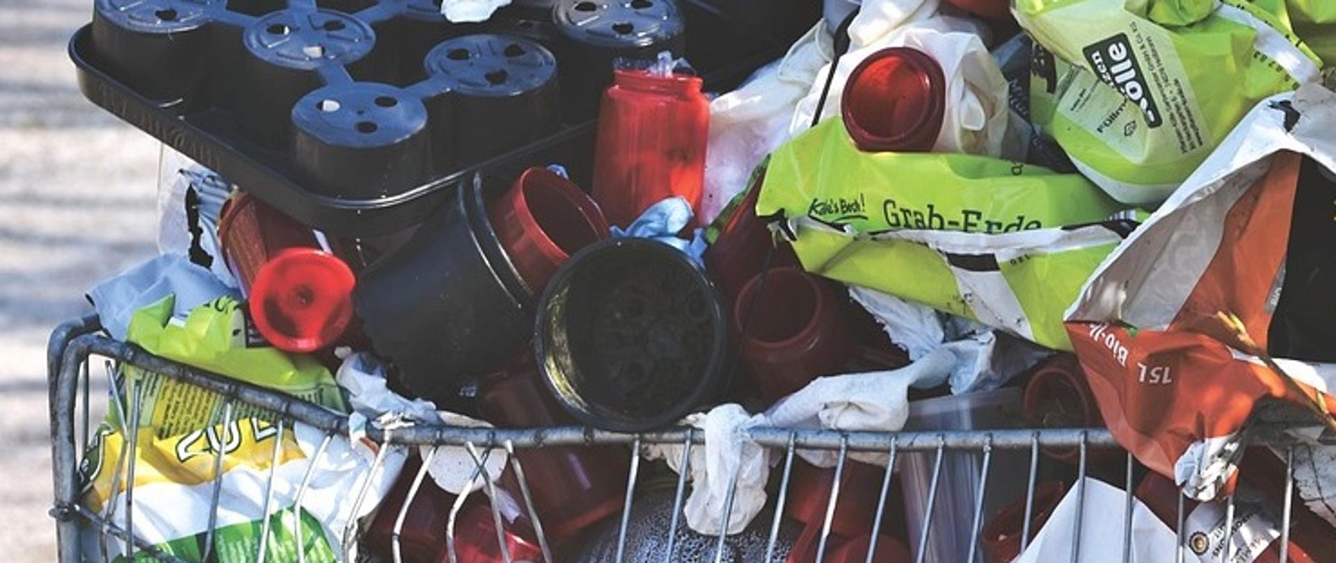 Mnóstwo różnych odpadów plastikowych i metalowych w dużym wózku sklepowym, stojącym na zewnątrz, na tle trawy