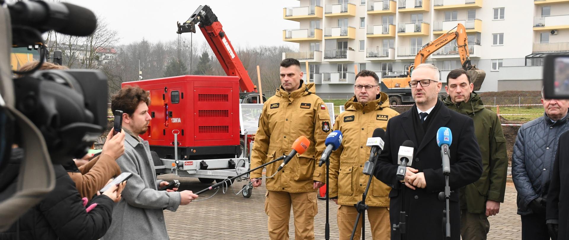 Przedstawiciele Państwowej straży pożarnej i politycy stoją otoczeni przez dziennikarzy na tle budowy. W tle widoczne koparki i zabudowania miejskie.