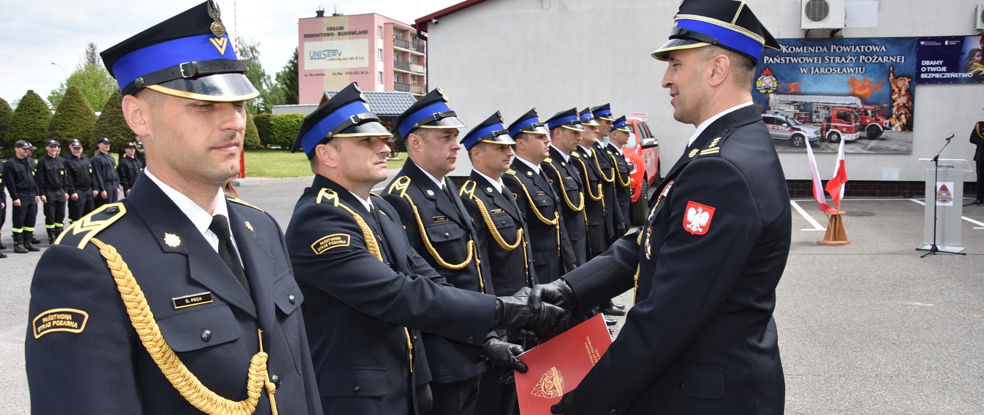 Zdjęcie zrobione na zewnątrz podczas uroczystości. Oficer Państwowej Straży Pożarnej w stopniu starszego brygadiera podaje dłoń Funkcjonariuszowi Państwowej Straży Pożarnej, gratulując otrzymanego wyróżnienia. 