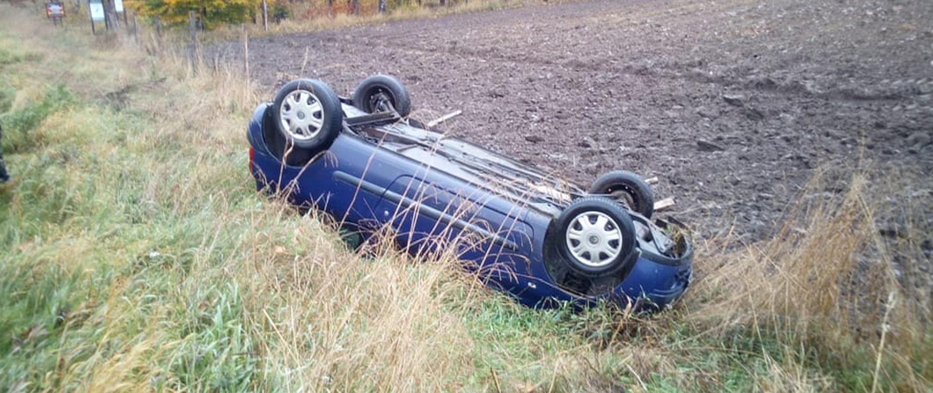 Niebieski samochód osobowy marki Renault Clio wypadł z drogi i leży na dachu na poboczu. Dookoła wysoka trawa, tereny leśne i pole.