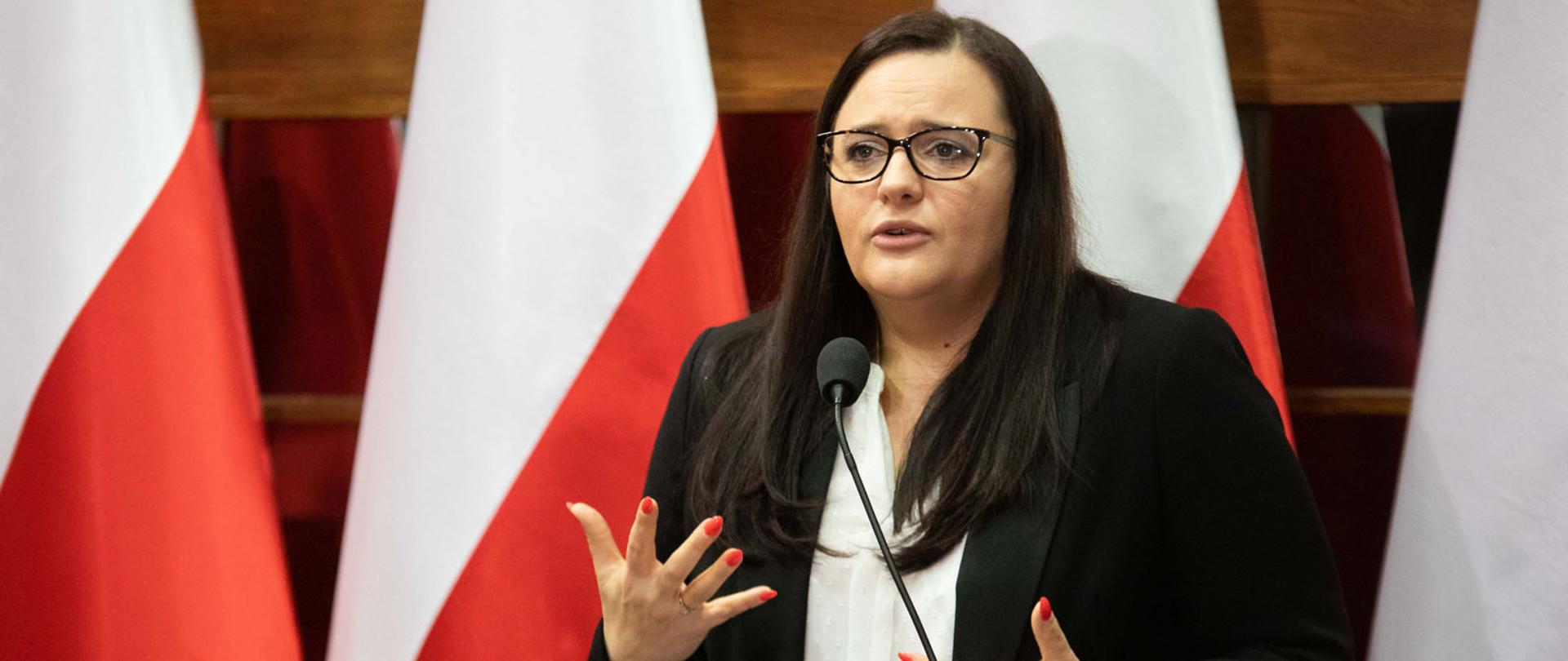 Na zdjęciu minister Małgorzata Jarosińska-Jedynak stoi przy mównicy, w tle cztery flagi biało-czerwone.
