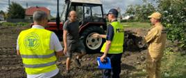 Na zdjęciu widać czerwony traktor marki Ursus stojący na polu. Obok niego znajdują się trzy osoby. Wszystkie osoby stoją na polu, jest ładna pogoda. Osoby rozmawiają ze sobą. W tle jest maszyny rolnicze i zielone pola.