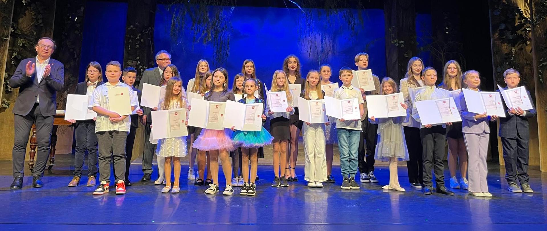 Niebieskie tło. Na zdjęciu stoi na scenie Lena Torba wraz z innymi osobami, wszyscy trzymają dyplomy.