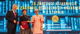 Na tle wielkiego ekranu z napisem Nagrody ministra stoi minister Wieczorek, wiceminister Mrówczyńska i kobieta w czarnej marynarce trzymająca czerwoną teczkę.