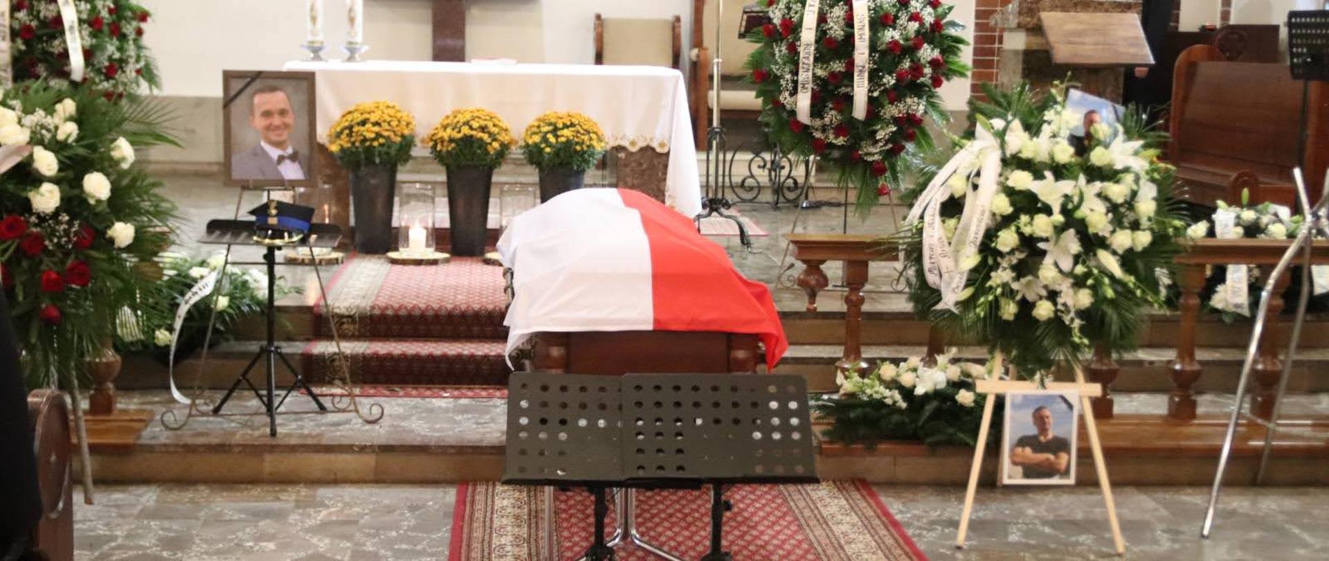 Trumna na której obłożona jest flagą Polski obok na sztalugach są wiązanki kwiatów po dwóch stronach trumny stoją zdjęcia zmarłego za trumną jest ołtarz oraz niedaleko stoi dwóch funkcjonariuszy Państwowej Straży Pożarnej.
