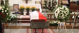 Trumna na której obłożona jest flagą Polski obok na sztalugach są wiązanki kwiatów po dwóch stronach trumny stoją zdjęcia zmarłego za trumną jest ołtarz oraz niedaleko stoi dwóch funkcjonariuszy Państwowej Straży Pożarnej.