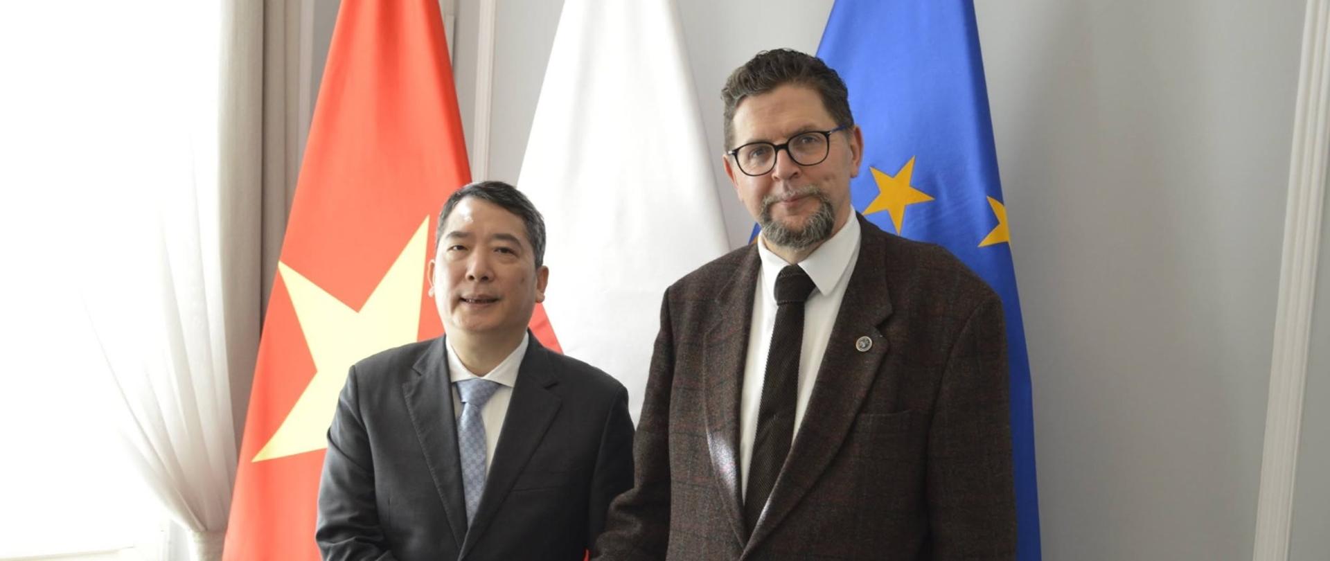 Pod białą ścianą stoją wiceminister Szeptycki i mężczyzna w czarnym garniturze, podają sobie ręce, za nimi flagi Polski, Wietnamu i UE, z lewej zasłonięte firanką okno.