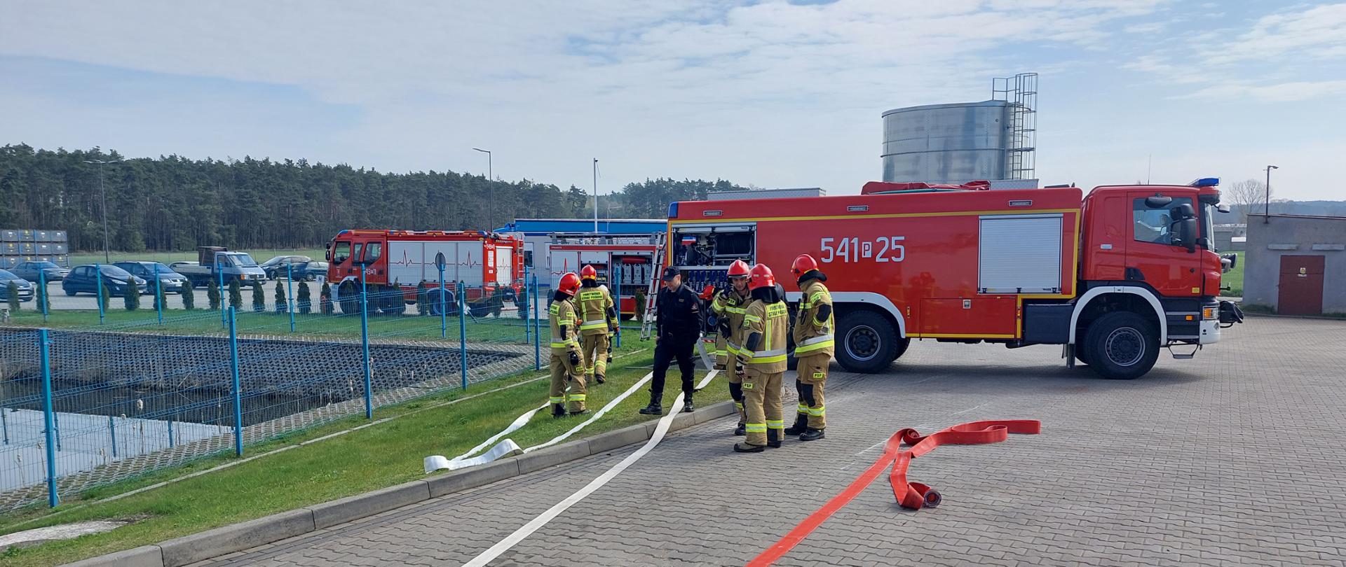 Strażacy w ubraniach specjalnych w trakcie rozwijania węży pożarniczych. Po lewej stronie zbiornik przeciwpożarowy, w tle wozy strażackie ustawione bokiem na terenie zakładu.